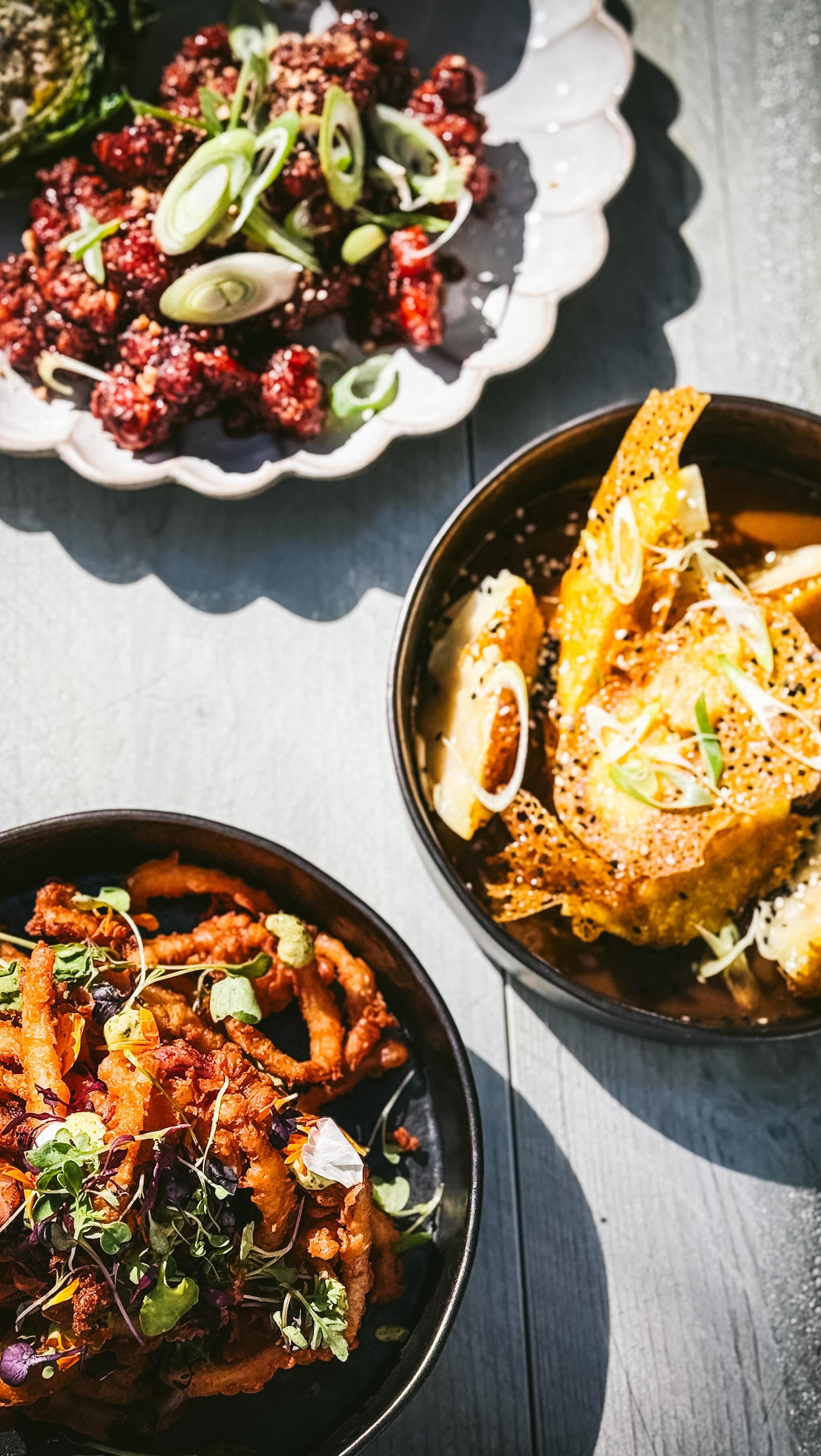 The image shows three dishes on a dark wooden surface: a plate of crispy fried items topped with green onions, a bowl of golden-brown tempura-style food garnished with sesame seeds and green onions, and another bowl containing fried onion rings with microgreens and edible flowers.