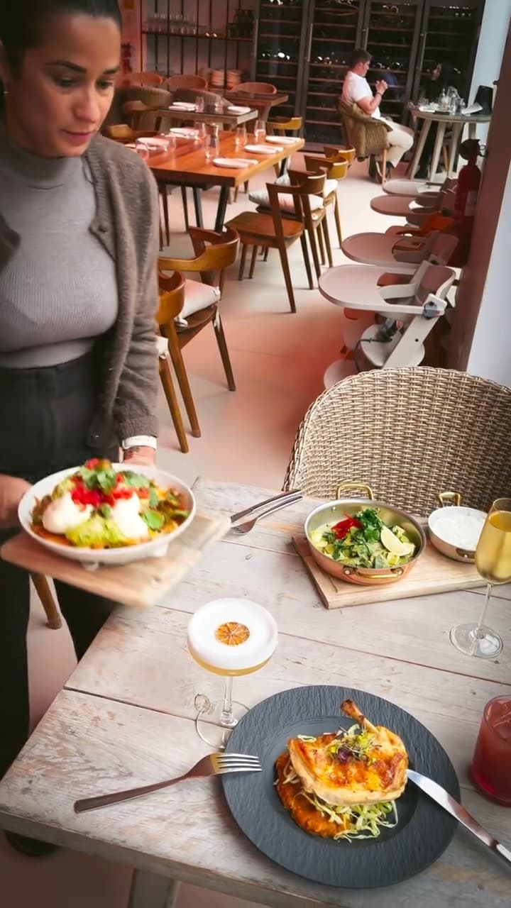 A server carries a bowl of salad topped with poached eggs and herbs toward a table set with a roasted chicken dish on a slate plate, a copper pan with a green salad, and two cocktails, one with a citrus garnish. The restaurant interior features wooden tables, wicker chairs, and a wine cellar in the background.