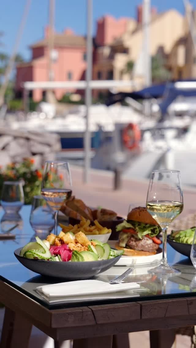 A table set outdoors by a marina features a burger with lettuce and tomato, a bowl of avocado and cucumber salad with fried elements, fries, and glasses of white and red wine. In the background, boats and waterfront buildings are visible under a clear sky.