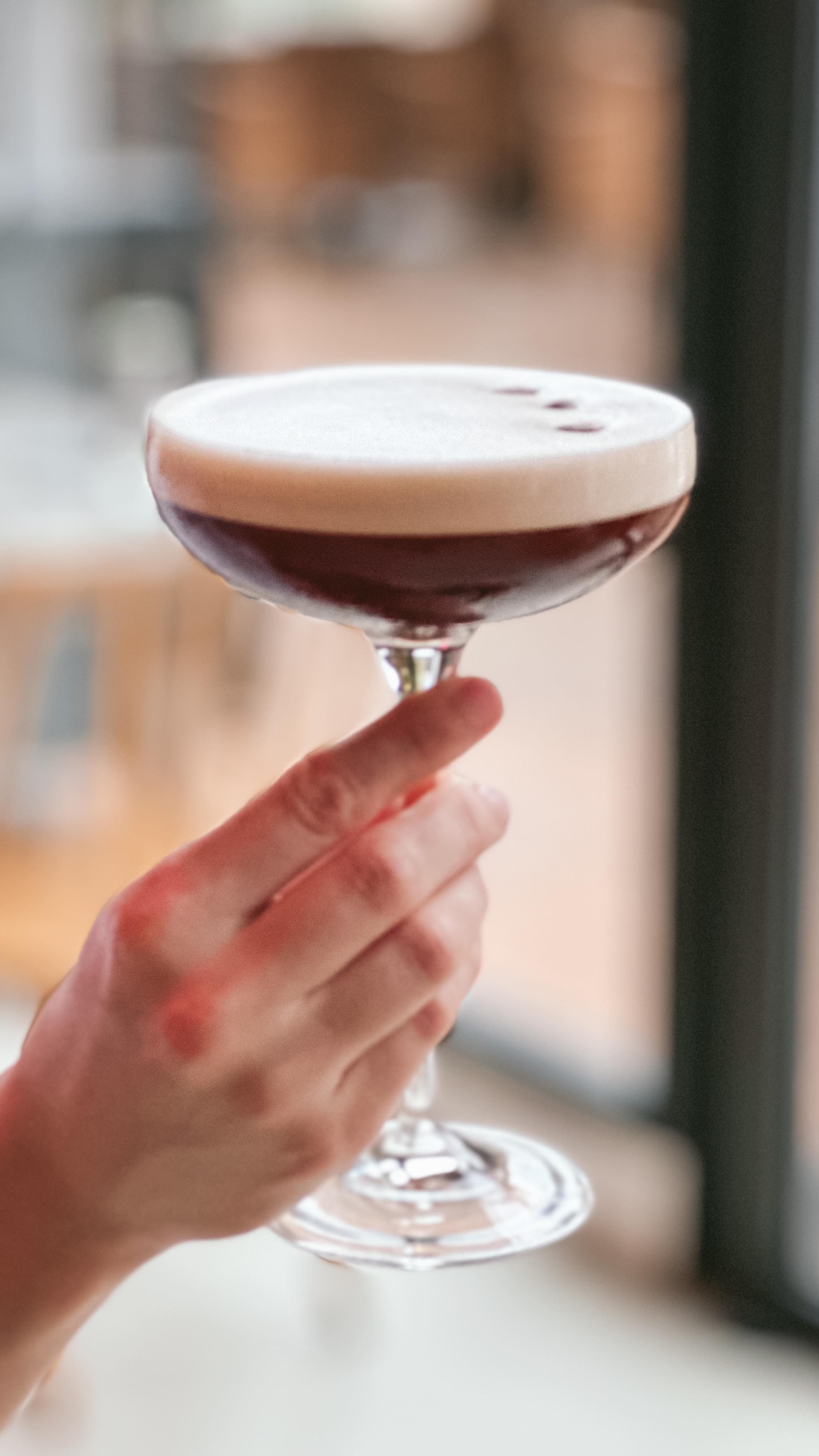 A hand holds a stemmed glass containing a dark red cocktail topped with a thick layer of white foam and garnished with small dark berries. The background is softly blurred, suggesting an indoor dining or bar setting.